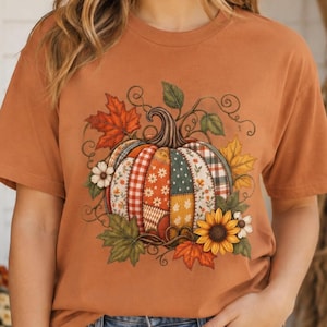 Puede incluir: Una camiseta de manga corta color naranja quemado con un gráfico central de una calabaza hecha de varios parches de tela estampada, incluyendo cuadros y flores, rodeada de hojas de otoño coloridas y girasoles amarillos.