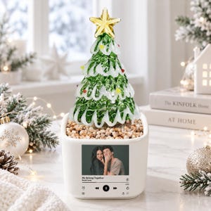 May include: A white square ceramic planter base featuring a printed music player interface with a photo of a couple and the text We Belong Together by Mariah Carey. A glass Christmas tree with green branches, white snow accents, and a yellow star topper.