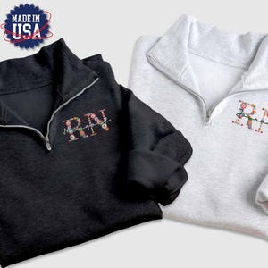 May include: Two quarter-zip sweatshirts, one black and one grey, with floral embroidered lettering. The black sweatshirt says "RN" and the grey sweatshirt says "RN".