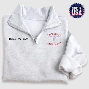 Puede incluir: Una sudadera gris con cremallera y un símbolo médico bordado en rojo con el texto "Emergency Department". El texto "Mason, RN, BSN" está bordado en negro debajo de la cremallera.
