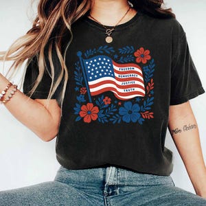 Op de afbeelding: Zwart T-shirt met een Amerikaanse vlag en de woorden "Vrijheid, Democratie, Gerechtigheid, Waarheid". De vlag is omgeven door rode en blauwe bloemmotieven. Het T-shirt is donker van kleur.