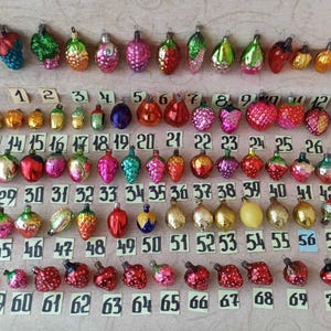 Könnte beinhalten: Eine Sammlung von siebzig weihnachtlichen Glasornamenten in verschiedenen Formen, darunter Früchte, Beeren und Zapfen, angeordnet in Reihen mit nummerierten Papieretiketten von 1 bis 70. Die Ornamente weisen diverse Farben wie Rot, Grün, Gold und Pink auf.