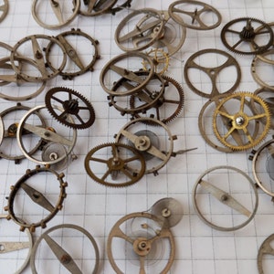 Puede incluir: Una colección de engranajes y muelles de relojes antiguos, algunos con diseños intrincados, dispuestos sobre una superficie blanca cuadriculada.