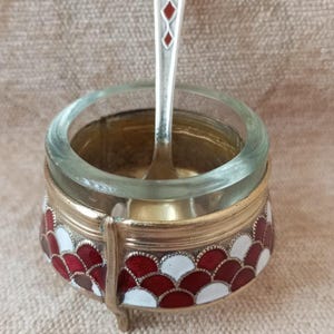 Puede incluir: Un salero vintage con un cuenco de cristal y una base metálica decorativa. La base está adornada con un patrón de esmalte rojo y blanco. Una pequeña cuchara con un mango decorativo descansa en su interior.