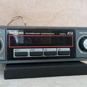 Puede incluir: Una radio de coche vintage con una placa frontal gris, con perillas para el volumen y la sintonización, y una pantalla digital. La radio tiene botones etiquetados en cirílico y la marca "Terrisa" es visible.
