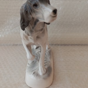 May include: A porcelain figurine of a grey and white hunting dog standing on a base with a textured surface.