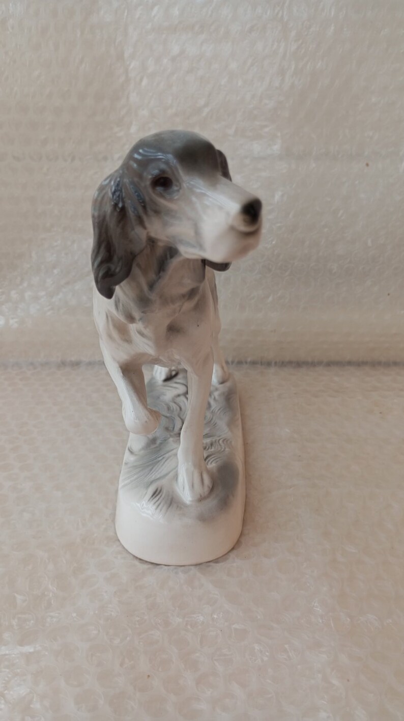 May include: A porcelain figurine of a grey and white hunting dog standing on a base with a textured surface.
