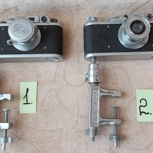 Puede incluir: Dos cámaras vintage plateadas con cubiertas de cuero negro están montadas en abrazaderas de metal plateado. Las cámaras están etiquetadas con "1." y "2."