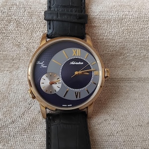 Puede incluir: Reloj de pulsera dorado con esfera azul oscuro y una esfera secundaria más pequeña. El reloj presenta agujas doradas y números romanos. La correa de cuero negro complementa el diseño elegante del reloj. La esfera del reloj tiene el texto "Dual Time" y "Adriatica".