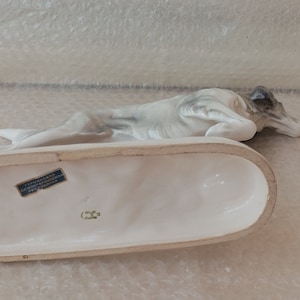 May include: A white porcelain figurine of a dog lying down on a white base. The base has a blue label with text that is partially visible. The dog's head is turned to the side and its eyes are closed.