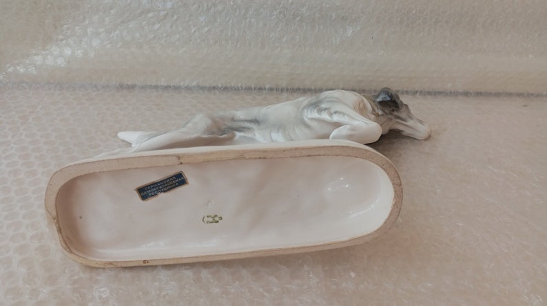 May include: A white porcelain figurine of a dog lying down on a white base. The base has a blue label with text that is partially visible. The dog's head is turned to the side and its eyes are closed.