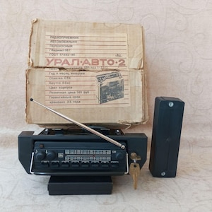May include: Vintage black car radio with a silver antenna, a key, and a separate black speaker. The radio is positioned in front of a cardboard box with Cyrillic text and the words "URAL-AVTO-2".