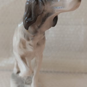 May include: A white porcelain figurine of a dog with black and grey markings. The dog is standing on a base with a grey and white design. The dog's head is tilted to the side and its eyes are closed.