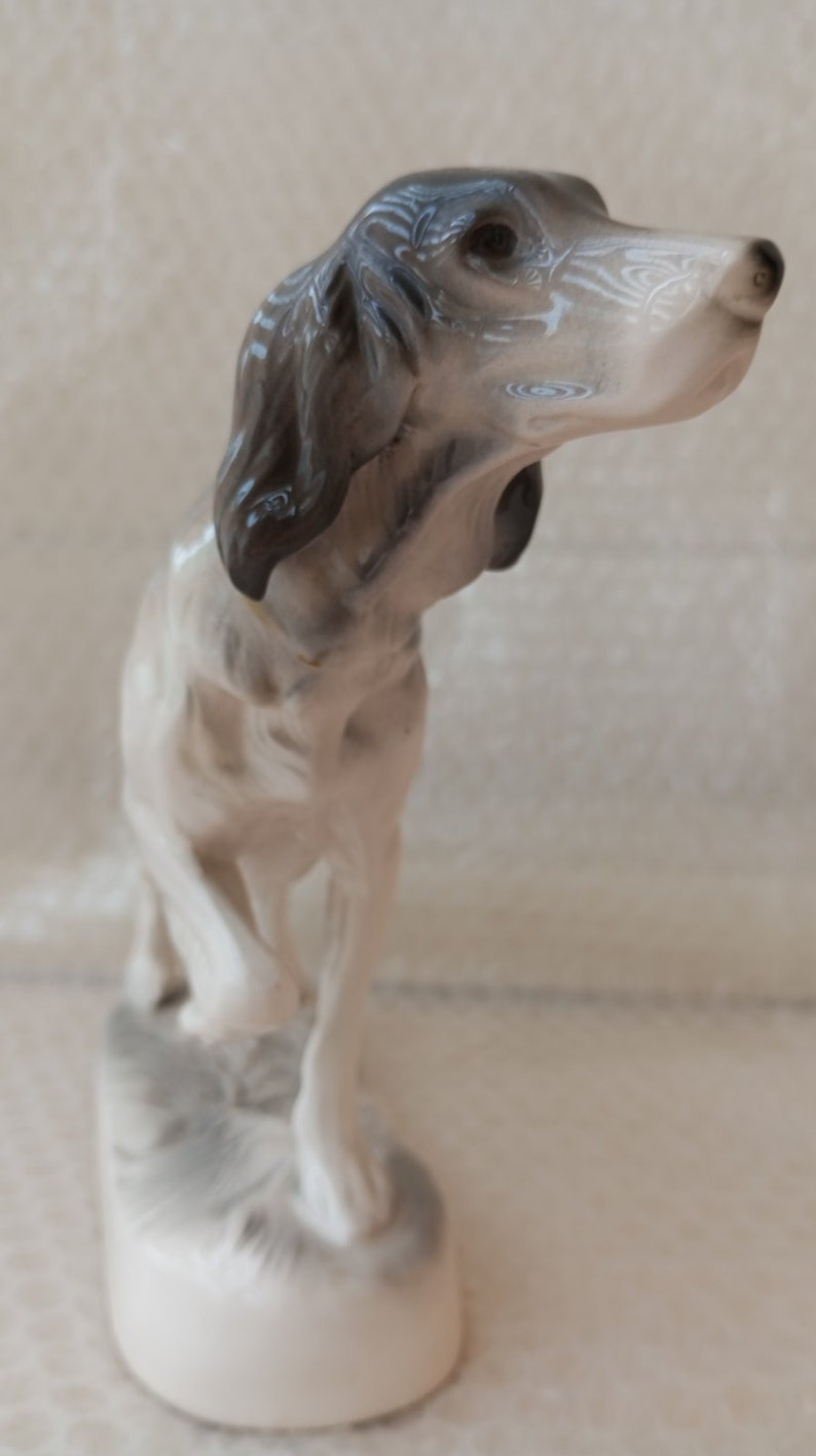 May include: A white porcelain figurine of a dog with black and grey markings. The dog is standing on a base with a grey and white design. The dog's head is tilted to the side and its eyes are closed.