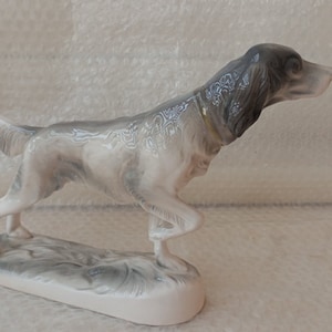 May include: A porcelain figurine of a grey and white hunting dog with a gold collar, standing on a base with a grey and white design.