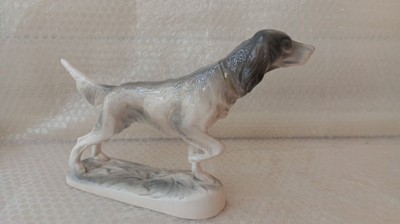 May include: A porcelain figurine of a grey and white hunting dog with a gold collar, standing on a base with a grey and white design.