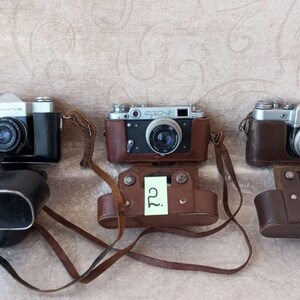 May include: Three vintage cameras with leather cases are displayed. The cameras have silver and black bodies with lenses. Each camera has a matching brown leather case and a neck strap. The cameras are numbered 1, 2, and 3.