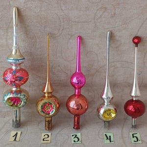 Op de afbeelding: Vijf vintage glazen kerstboompieken op een rij, genummerd van één tot vijf. De ornamenten hebben diverse vormen, waaronder bollen en langwerpige punten, in metallic kleuren zoals rood, goud, zilver en roze met geschilderde bloemmotieven.