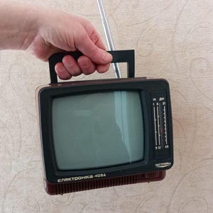 May include: A vintage portable television with a black and maroon casing, a handle, and an extendable antenna. The screen is gray, and the side features a tuning dial. The text "Электроника-404Д" is visible.