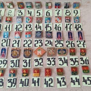 May include: A collection of vintage enamel pins and badges, each with a unique design and color scheme. The pins are arranged on numbered cards, creating a display of collectible items. The numbers are written in black ink.