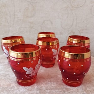 Op de afbeelding: Zes rode glazen bekers met gouden randen en witte bloemen- en stippenontwerpen. De glazen hebben een afgeronde vorm en zijn gerangschikt op een lichtgekleurd oppervlak. De randen en de basis van de glazen zijn goudkleurig.