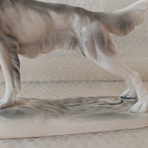 May include: A white porcelain figurine of a dog running, with a grey base. The dog is in a mid-stride pose, with its tail raised and its head turned to the side.