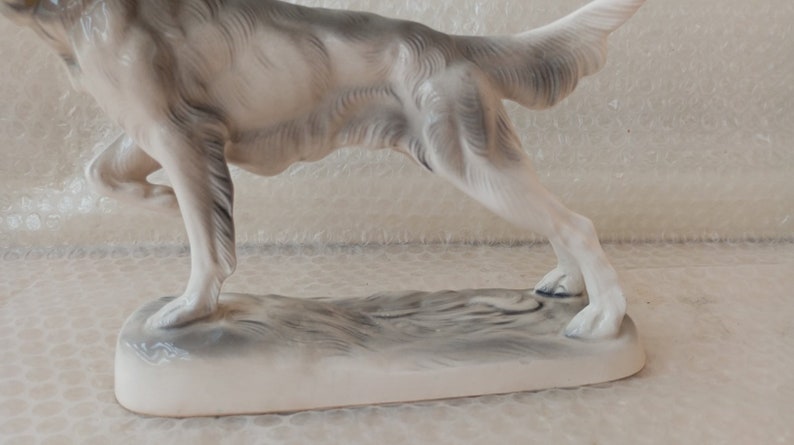 May include: A white porcelain figurine of a dog running, with a grey base. The dog is in a mid-stride pose, with its tail raised and its head turned to the side.