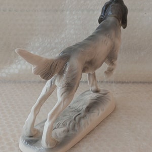 May include: A porcelain figurine of a grey and white hunting dog with its head turned back, standing on a base with a grey and white design.