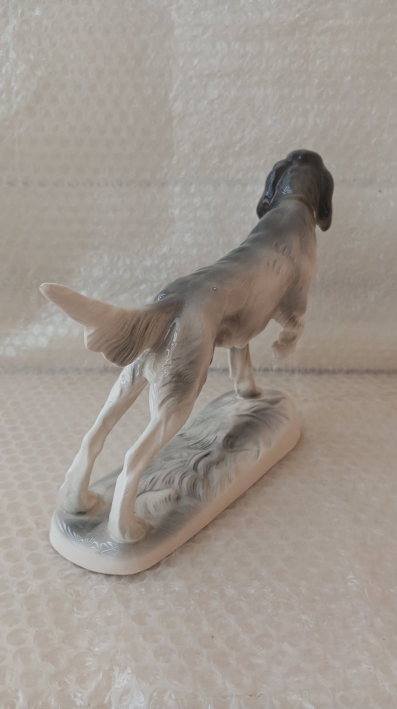 May include: A porcelain figurine of a grey and white hunting dog with its head turned back, standing on a base with a grey and white design.