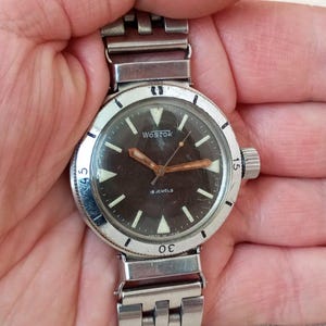 May include: A vintage wristwatch with a silver metal strap and case. The watch face is dark with luminous markers and hands. The word "VOSTOK" is printed on the face. The watch has a rotating bezel and a crown on the side.