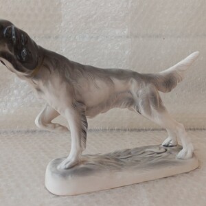 May include: A porcelain figurine of a grey and white hunting dog with a black nose and ears. The dog is standing on a white base with a grey and white textured surface.