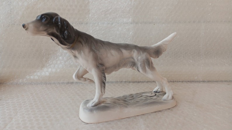 May include: A porcelain figurine of a grey and white hunting dog with a black nose and ears. The dog is standing on a white base with a grey and white textured surface.