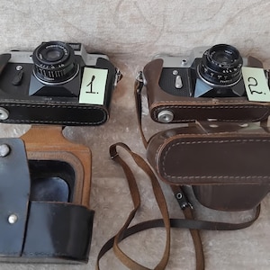 May include: Two vintage cameras with leather cases. The cameras are black and silver with a brown leather case. The cameras are labeled with numbers 1 and 2.