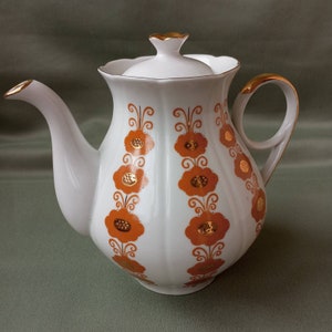 Könnte beinhalten: Weiße Keramik-Teekanne mit Deckel, verziert mit orangefarbenen und goldenen Blumenmustern. Die Teekanne hat einen gebogenen Ausguss und Griff, mit goldenen Verzierungen an Ausguss, Griff und Deckel. Das Blumenmuster ist in vertikalen Reihen angeordnet.