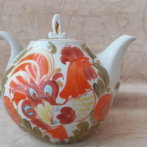 Könnte beinhalten: Weiße Keramik-Teekanne mit einem lebendigen Hahn-Design. Die Teekanne hat einen gebogenen Griff, einen Ausguss und einen Deckel mit dekorativem Knauf. Der Hahn ist in Rot-, Orange- und Blautönen mit floralen Akzenten dargestellt.