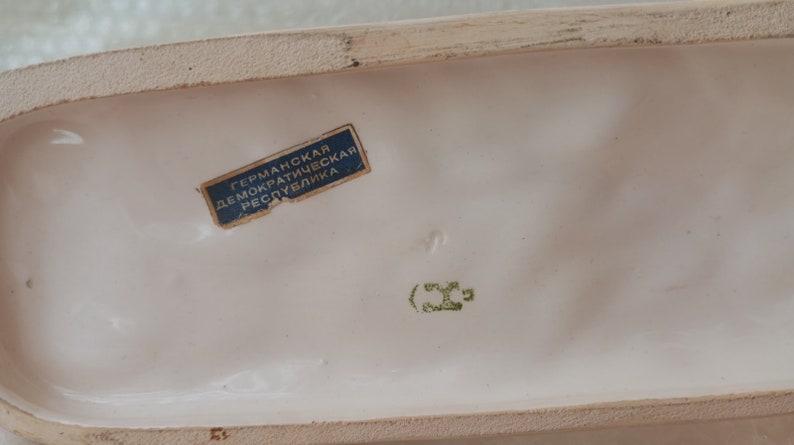 May include: A white ceramic figurine with a blue label that reads "German Democratic Republic". The figurine has a green mark that reads "(36".