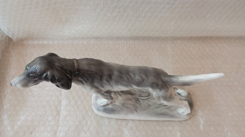 May include: A porcelain figurine of a greyhound dog running, with a white tail and base. The dog is in a hunting pose, with its head turned to the side.