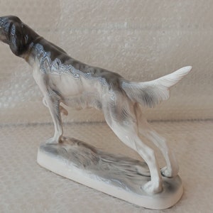May include: A porcelain figurine of a grey and white hunting dog standing on a base. The dog is posed in a running position with its head turned to the side.