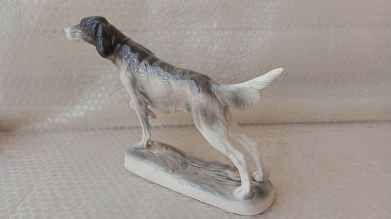 May include: A porcelain figurine of a grey and white hunting dog standing on a base. The dog is posed in a running position with its head turned to the side.
