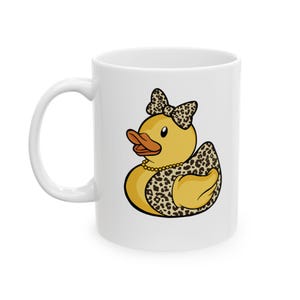 Könnte beinhalten: Eine weiße Keramiktasse mit schwarzem Henkel und dem Text "But First, Coffee" in fetter, schwarzer Schrift. Die Tasse steht vor einem verschwommenen Hintergrund, der eine gemütliche Umgebung suggeriert.