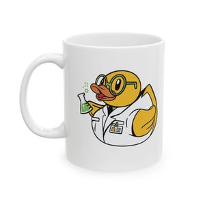 Könnte beinhalten: Weiße Keramik-Tasse mit einem Cartoon-Enten-Design, die eine Brille und einen Laborkittel trägt und einen Kolben mit grüner Flüssigkeit hält. Die Ente hat einen orangefarbenen Schnabel und ein Namensschild. Die Tasse hat einen weißen Griff.