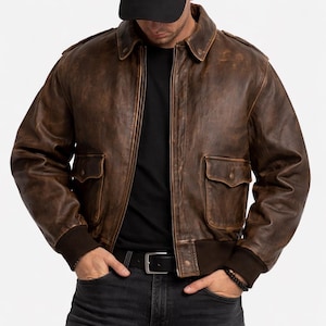 Könnte beinhalten: Braune Leder-Bomberjacke mit zwei Vordertaschen und geripptem Saum. Die Jacke hat ein Used-Finish und einen klassischen Kragen. Die Person im Bild trägt eine schwarze Kappe und eine schwarze Jeans.