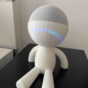 Puede incluir: Soporte blanco impreso en 3D con forma de persona con cabeza redonda y miembros cortos. El soporte está diseñado para sostener un altavoz inteligente.