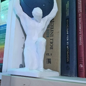 May include: A white 3D printed sculpture of a person holding a dark grey round speaker. The sculpture is on a white base and is sitting on a bookshelf with several books. The books have titles in Romanian and French.