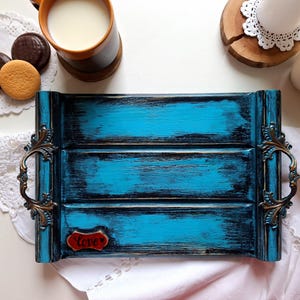 May include: A rectangular wooden serving tray featuring a distressed turquoise and black finish with ornate bronze-toned metal handles on each side. A small red plaque with the word "love" in black script is attached to the bottom left corner.