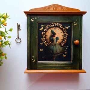 Könnte beinhalten: Ein grüner Schlüsselkasten aus Holz mit einem goldenen Zifferblatt und einer Frau in einem grünen Kleid. Das Zifferblatt ist mit goldenen Schmetterlingen verziert. Der Schlüsselkasten hat einen kleinen Holzknopf an der Tür.
