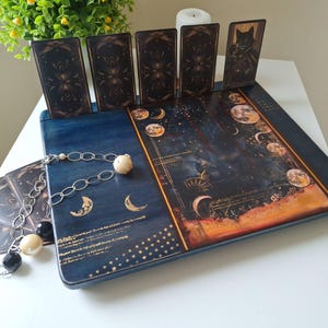Könnte beinhalten: Ein dunkelblauer Tarotkartenhalter aus Holz mit einem Himmelsdesign, das Monde, Sterne und ein Nachthimmelmotiv zeigt. Der Halter zeigt mehrere Tarotkarten mit verzierten Rückseiten. Eine dekorative Kette mit Perlen und Anhängern liegt auf dem Halter.
