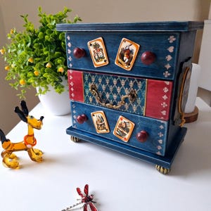 May include: A small, blue wooden chest of drawers with three drawers, decorated with playing card images. The drawers have red and patterned accents, and dark red knobs. The chest is decorated with playing card images and decorative accents.