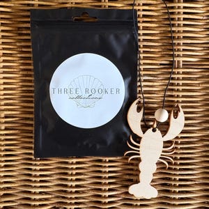 May include: A wooden lobster-shaped pendant with a black cord, next to a black pouch with a white label. The label has the text "THREE ROOKER collections" and a seashell graphic. The background is a woven wicker pattern.