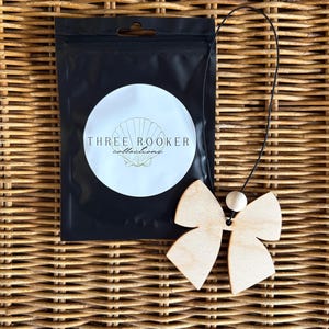 May include: A light-colored wooden bow ornament featuring a small round wooden bead and a black string for hanging, positioned next to a black plastic pouch with a white circular label displaying a shell graphic and the text THREE ROOKER collections.